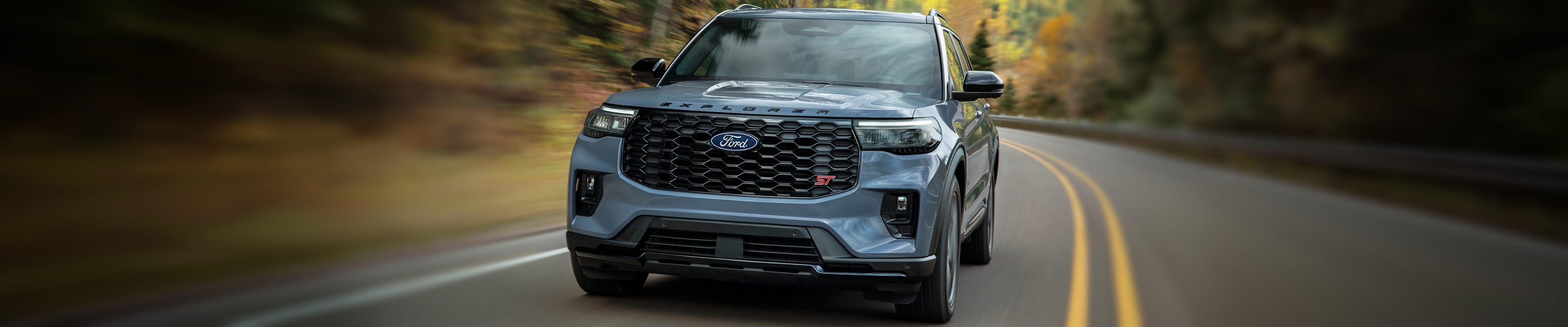 Blue-gray Ford Explorer ST SUV driving along a winding road, shown from the front with a black grille and sporty styling.
