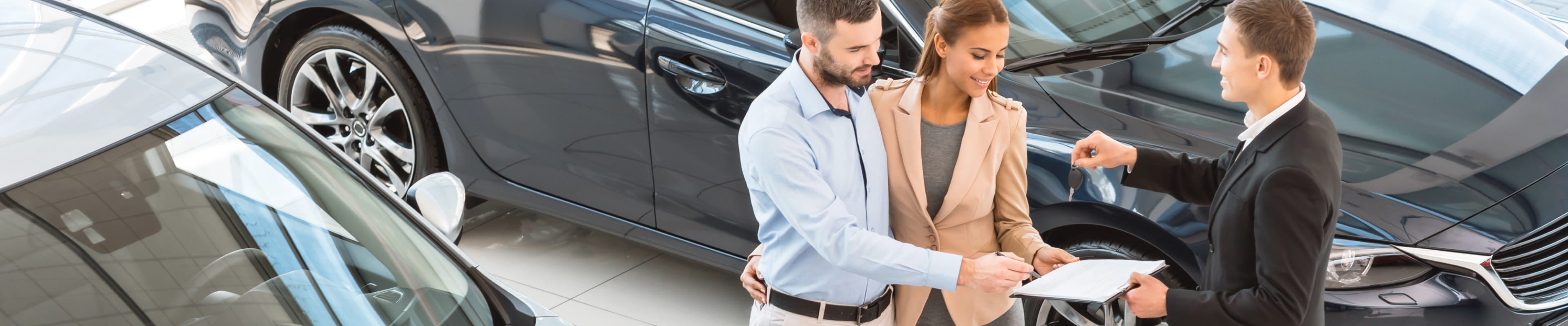 Car salesperson handing vehicle keys to a couple while they review and sign paperwork inside a dealership showroom.